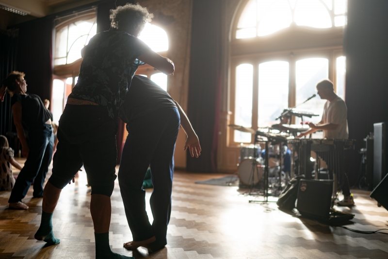 Foto van dansend persoon met twee muzikanten op de achtergrond, één muzikant met een drumstel en één met keyboard en microfoon,, beeld van deze Open Dance Jam