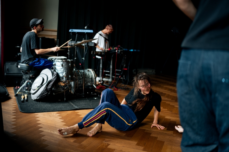 Foto van dansend persoon met twee muzikanten op de achtergrond, één muzikant met een drumstel en één met keyboard en microfoon,, beeld van deze Open Dance Jam